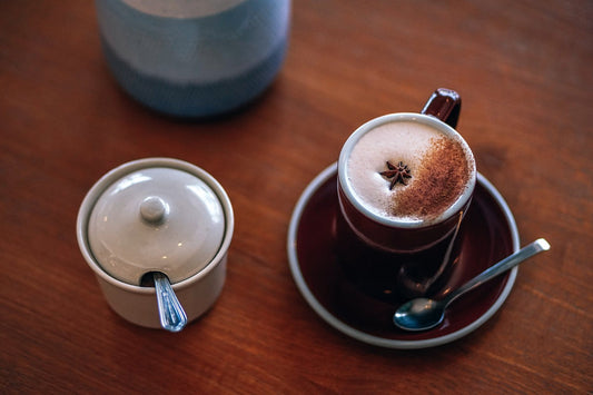 a photo of a cup of chai with milk and spices on top and a covered bowl of sugar to the left.