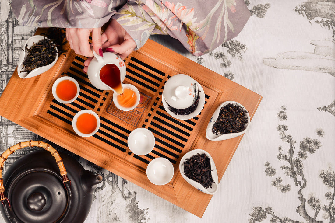 Image of a gong fu cha ceremony with a light brown tea tray upon which is setting white cups, tea pots, containers of dry tea and someone is pouring tea from a tea pitcher into the cups